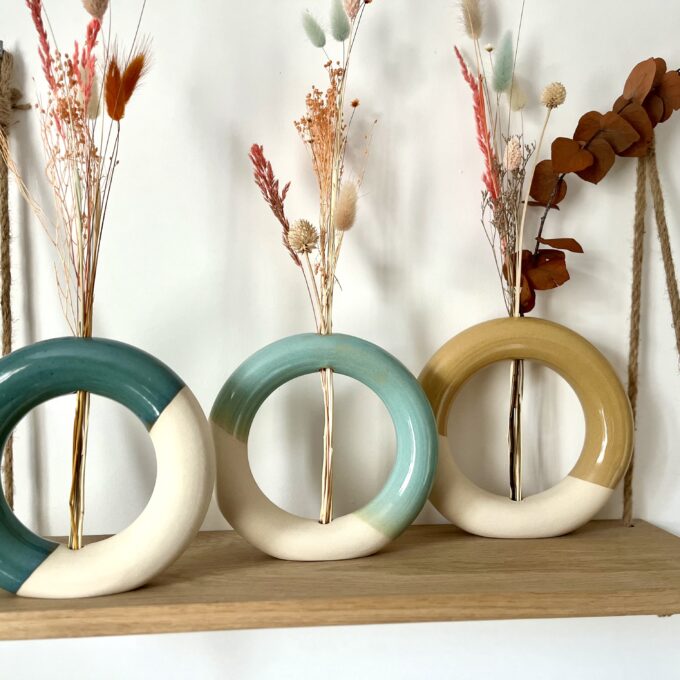 Trois vases artisanaux en céramique en forme donuts posés sur une étagère avec des fleurs séchées