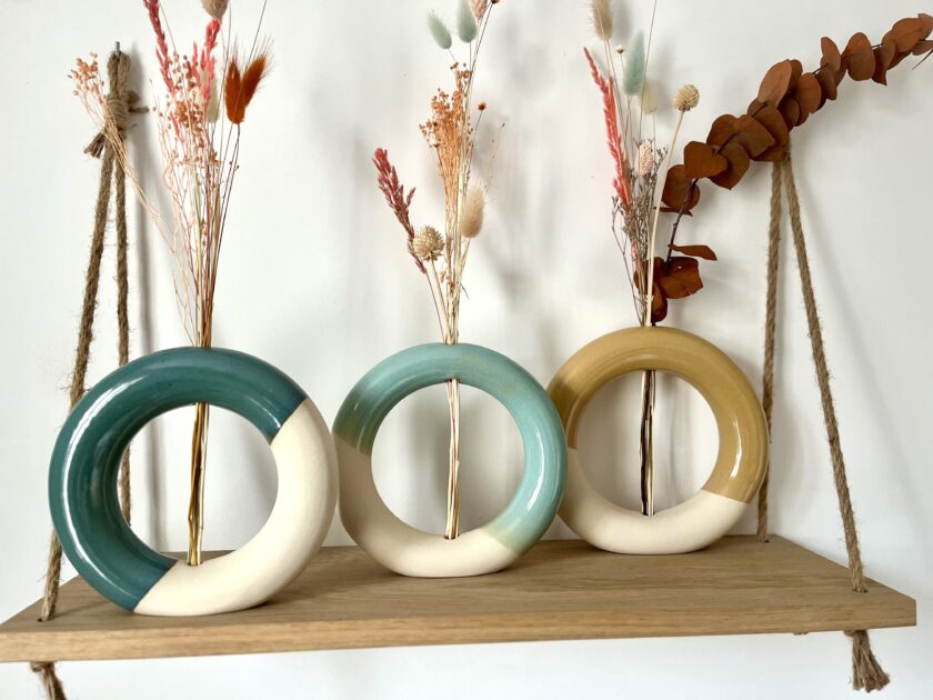 Trois vases artisanaux en céramique en forme donuts posés sur une étagère avec des fleurs séchées